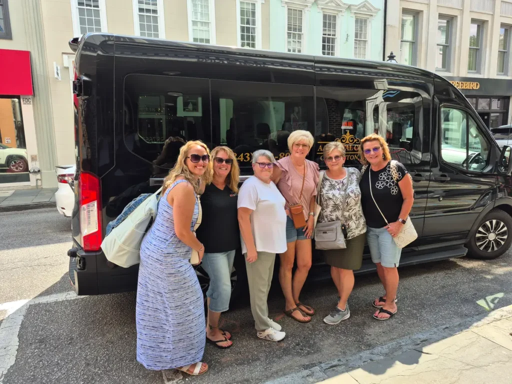 A group of smiling women passengers posing outside a black AAA Royal Coach passenger van on a historic street in downtown Charleston, SC.